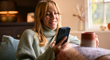 A smiling person is sitting on a couch with a cup of coffee in one hand and a smart phone in the other.