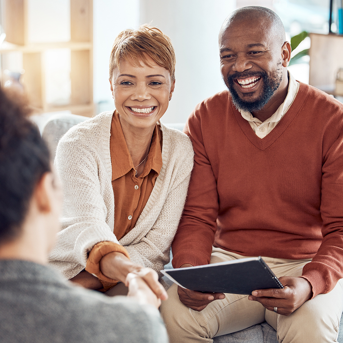 A man and woman are sitting on a couch. The man is holding an informational brochure in his hands and the woman is shaking heads with another woman who is sitting across from them. You just see the back of her head.