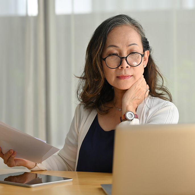 Woman wearing black glasses is sitting at a desk and looking at a laptop screen. She’s holding papers in her hand and there’s a tablet on the table in front of her.