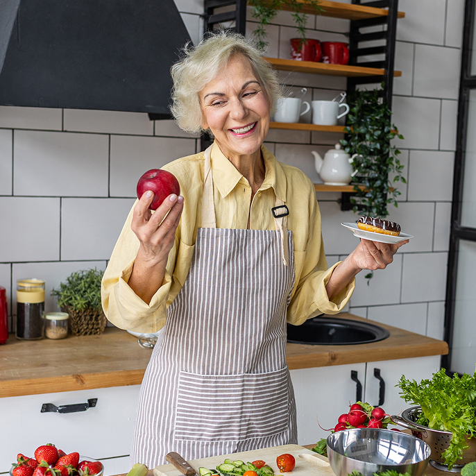 Older woman is standing in a kitchen. She has an apple in one hand and a donut on a plate in the other. She’s looking at the hand with the apple.