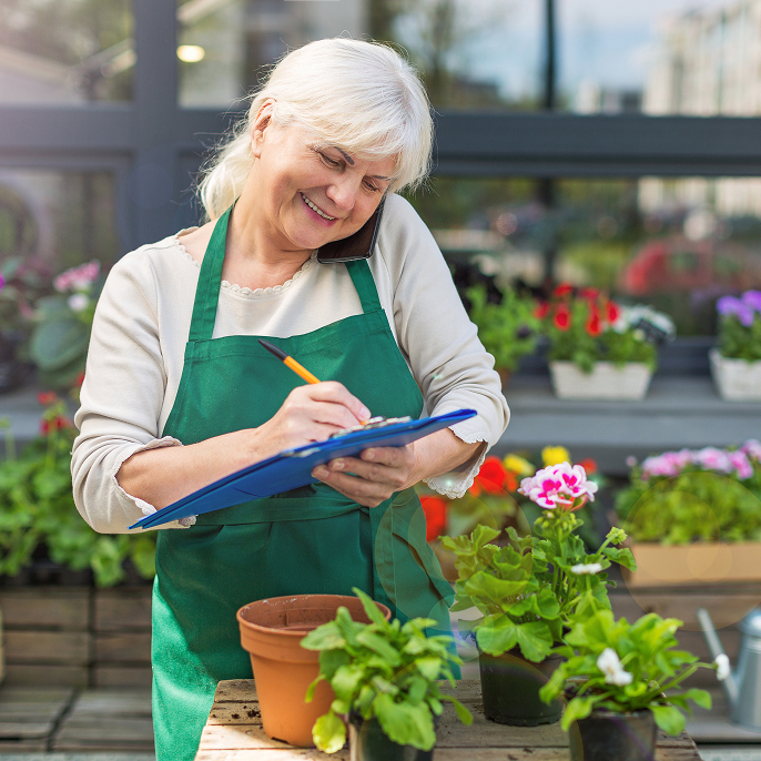 Older woman working in a greenhouse. She’s wearing a green apron and is holding a clipboard and a pen while holding a cellphone between her ear and her shoulder.