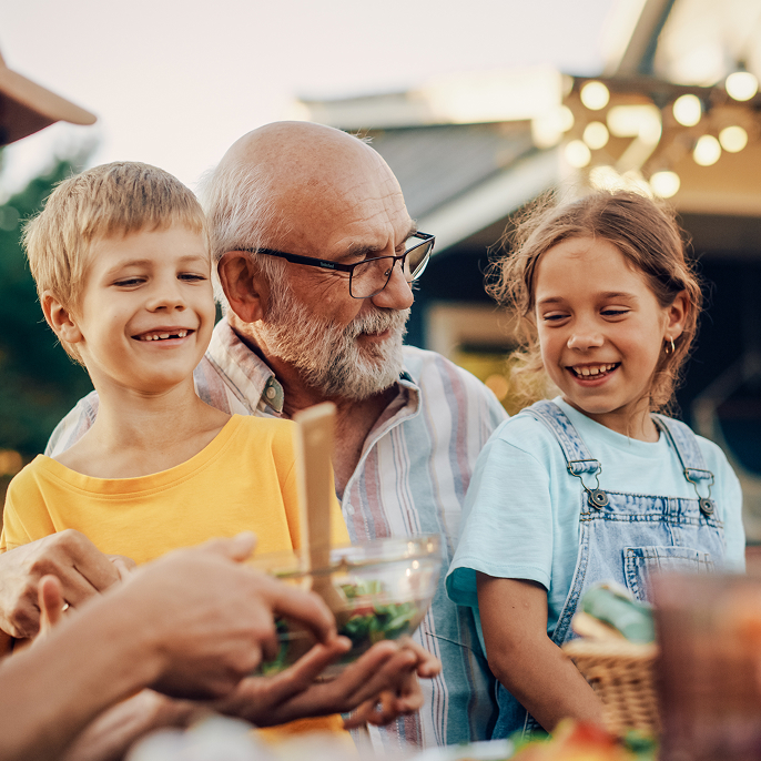 Grandfather sitting with his two young grandchildren on his lap. They’re outside sitting at a table where food is being passed around in the foreground.