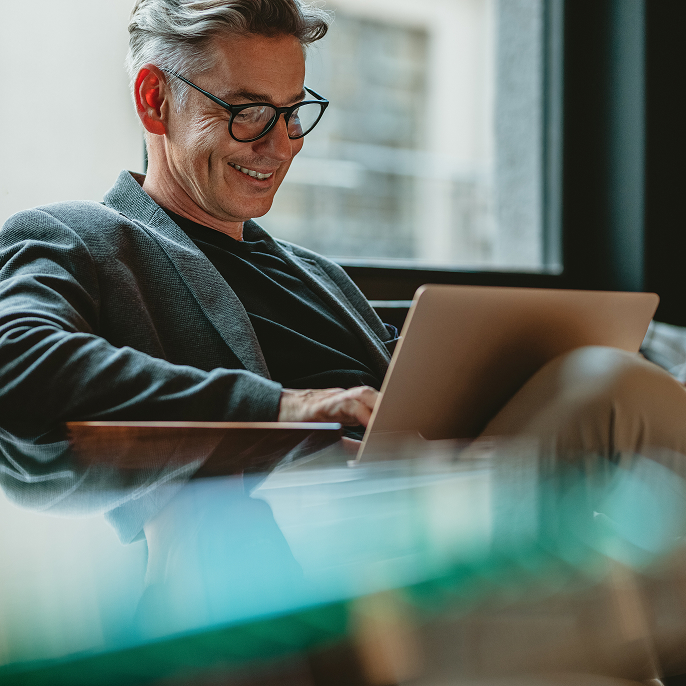 Middle aged man wearing glasses sitting in a chair in front of a window. He has a laptop on his lap