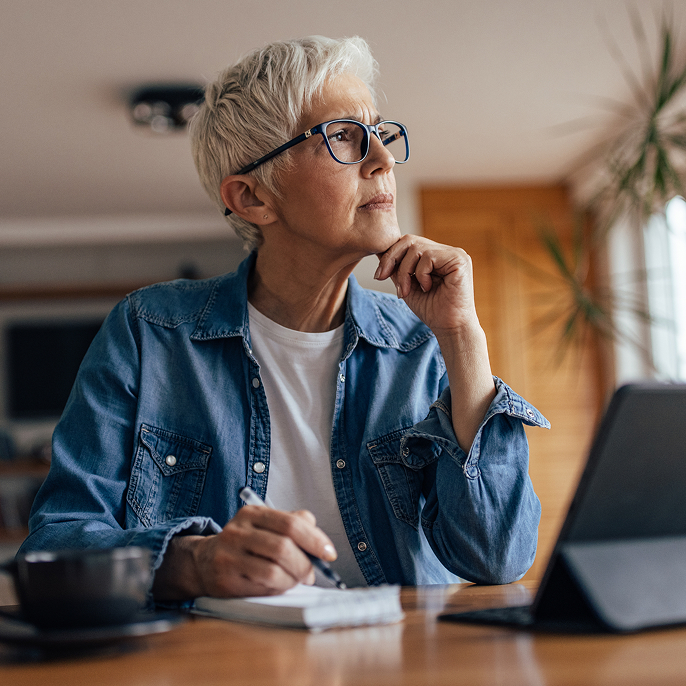 Older woman wearing black-framed glasses is looking off in the distance. She’s holding a pen and has a notebook and a tablet in front of her.