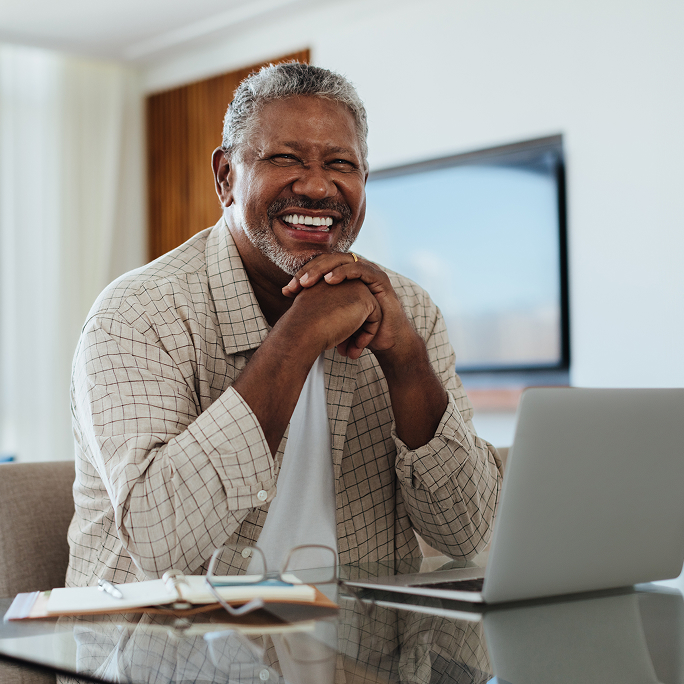 Smiling man sitting at a table. He’s looking at the camera and there’s a laptop on the table in front of him.