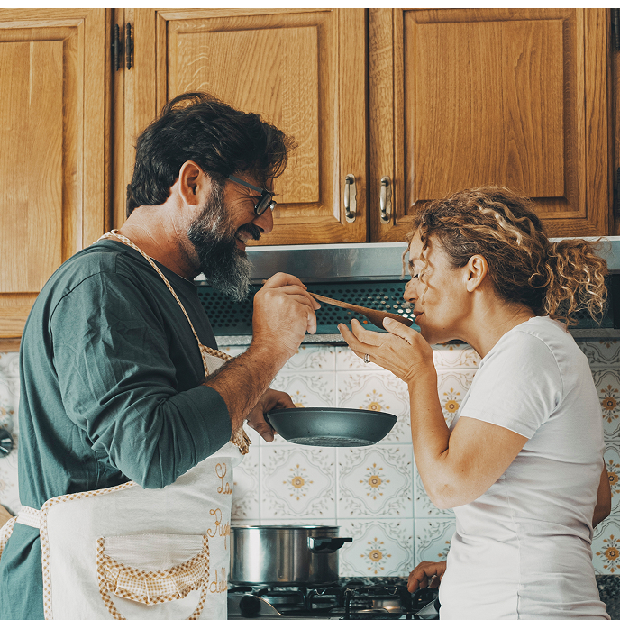 Man wearing an apron is holding a skillet and feeding a woman with a wooden cooking spoon.